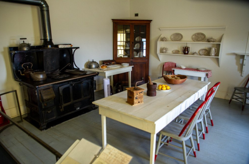 A vintage kitchen featuring a black stove, wooden table, chairs, and shelves with dishes and a bowl of oranges.