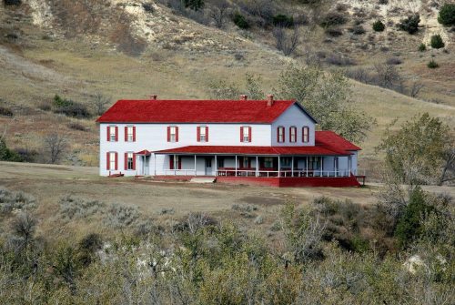 A large two-story house with a red roof, surrounded by rolling hills and sparse vegetation.
