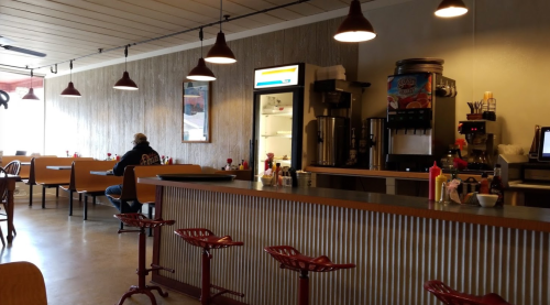 A cozy diner interior with a counter, stools, and a customer sitting alone at a table. Soft lighting and decor create a welcoming vibe.