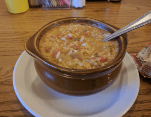 A bowl of soup with rice and tomatoes, served on a white plate, with a spoon resting inside.