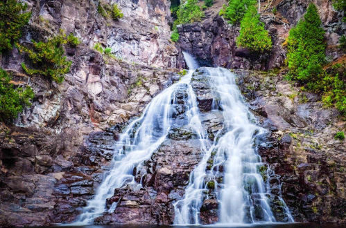 A cascading waterfall flows down rocky cliffs, surrounded by lush green trees and vibrant natural scenery.