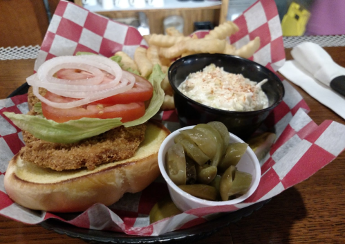 A plate with a fried chicken sandwich topped with lettuce, tomato, and onions, served with coleslaw, pickles, and fries.