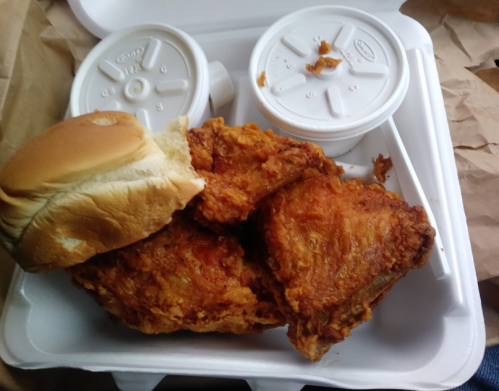 A takeout container with fried chicken pieces, a bun, and two cups, likely containing sides or sauces.