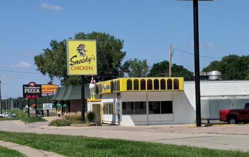 A roadside view of Sneaky's Chicken restaurant with a bright yellow sign, surrounded by trees and nearby businesses.