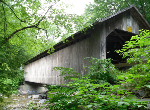 A wooden covered bridge spans a small creek, surrounded by lush green trees and foliage.