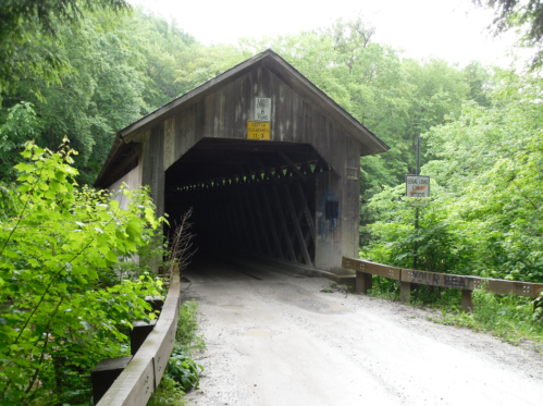 A wooden covered bridge surrounded by lush green trees, leading into a dark tunnel-like entrance.
