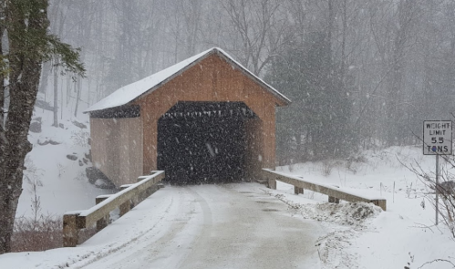 A snow-covered road leads to a wooden covered bridge in a winter landscape, with heavy snowfall and trees in the background.