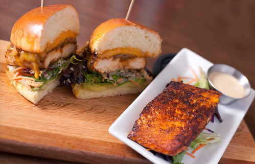 A wooden platter featuring two chicken sandwiches with melted cheese and a side of grilled fish with salad.