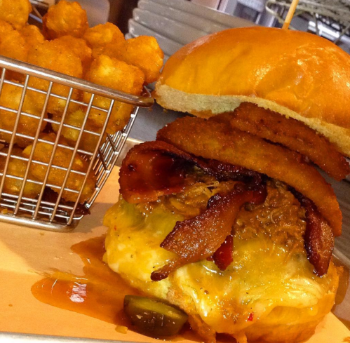 A juicy burger topped with bacon and onion rings, served with a side of crispy tater tots in a small basket.