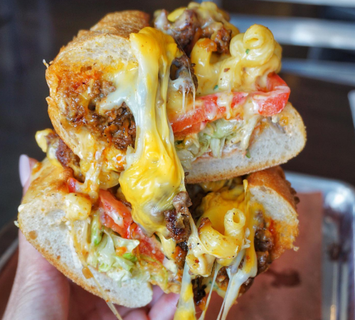A close-up of a cheesy sandwich filled with ground beef, macaroni, lettuce, and tomatoes, held in a hand.