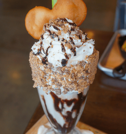 A tall glass of ice cream topped with whipped cream, chocolate drizzle, crushed cookies, and a cookie shaped like a Mickey Mouse head.