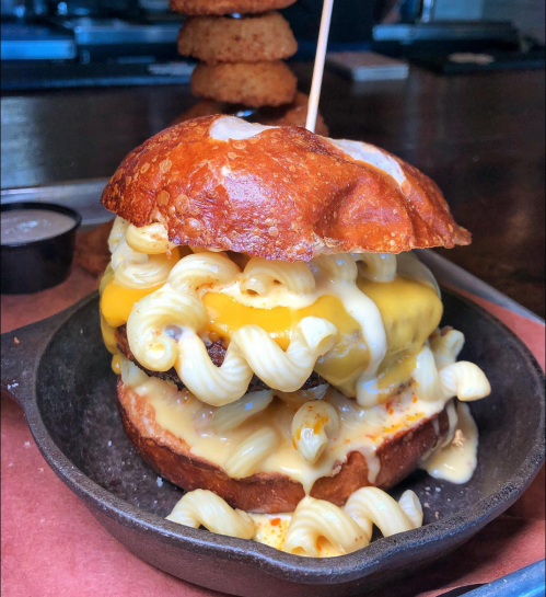 A hearty burger with melted cheese and macaroni piled high, served on a toasted bun in a cast iron skillet.