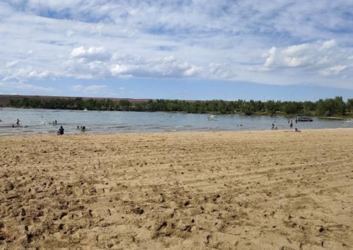 A sandy beach by a river with people swimming and enjoying the water under a partly cloudy sky.