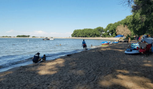 A sandy beach by a calm lake, with people enjoying the water and boats in the distance under a clear blue sky.