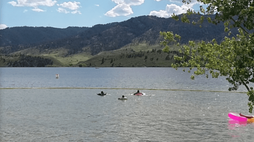 Two people swim in a calm lake surrounded by green hills and blue skies with fluffy clouds.