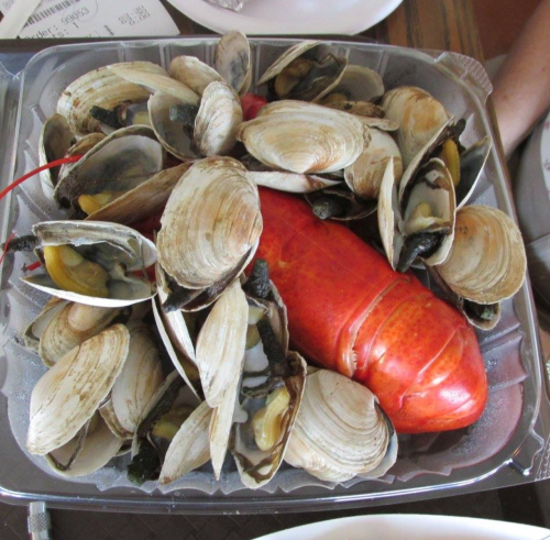 A platter of seafood featuring a large red lobster surrounded by clams in a clear container.