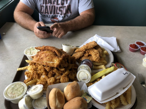A large tray of fried food, including chicken, shrimp, fries, and rolls, with a person using a phone in the background.