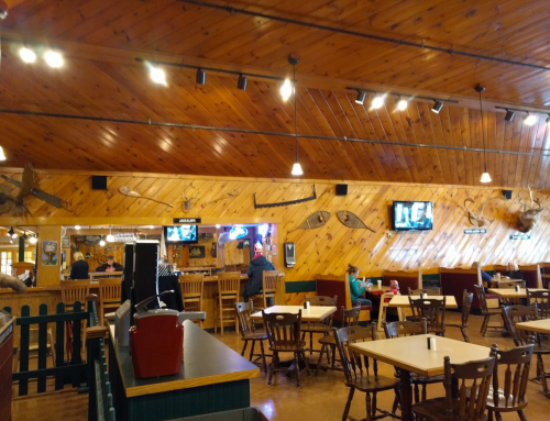 Interior of a rustic restaurant with wooden walls, tables, and a bar area, featuring TVs and decorative fishing gear.
