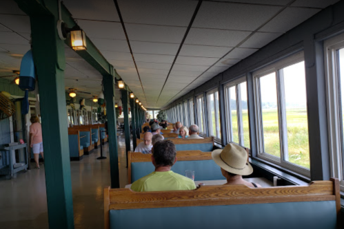 A spacious dining area with patrons seated in booths, large windows offering views of a grassy landscape outside.