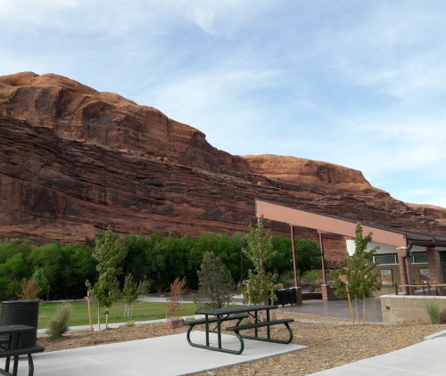 A picnic area with tables in front of a towering red rock formation and green trees under a blue sky.