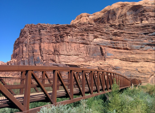A metal bridge spans a green area, set against towering red rock formations under a clear blue sky.
