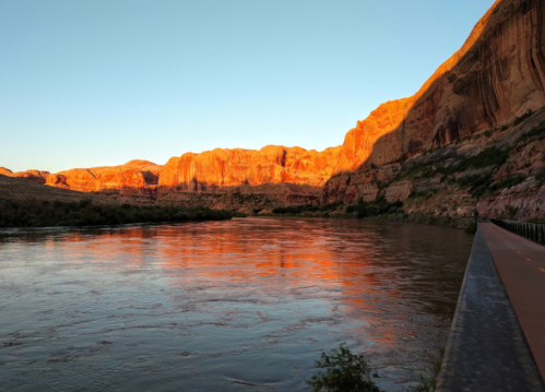 Sunset over a river, reflecting orange hues on the water, with rocky cliffs in the background.