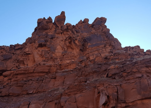A rugged red rock formation rises against a clear blue sky, showcasing jagged peaks and textured layers.