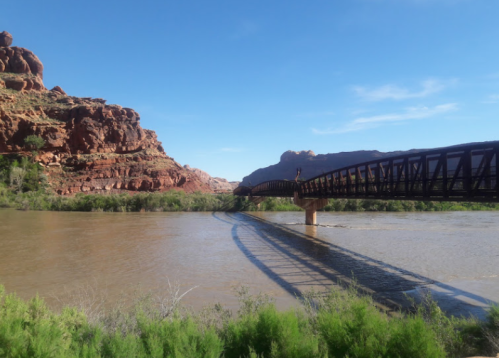 A wooden bridge spans a river, surrounded by red rock formations and greenery under a clear blue sky.
