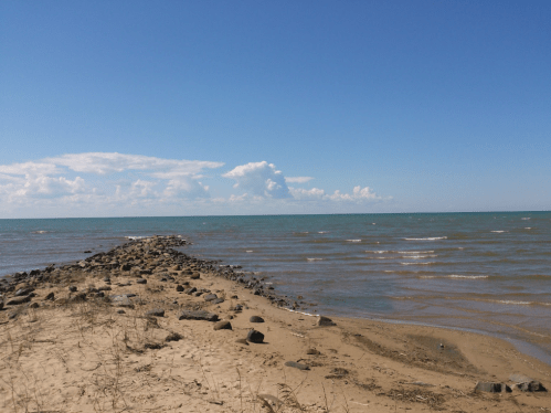 A sandy shoreline leads to a rocky jetty extending into a calm blue lake under a clear sky with scattered clouds.