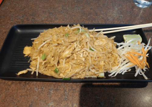 A plate of pad thai with chicken, bean sprouts, lime, and carrots, served with chopsticks on a black tray.