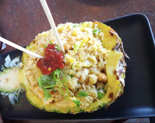 Fried rice served in a hollowed pineapple, garnished with herbs and a spicy sauce, with chopsticks for serving.