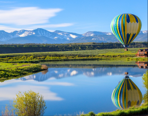 A colorful hot air balloon floats above a calm lake, reflecting mountains and blue skies in the background.