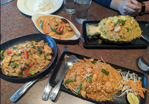 A table with three dishes: pad thai, stir-fried noodles, and a seafood rice dish, alongside rice and water glasses.