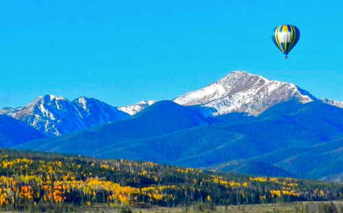 A colorful hot air balloon floats over snow-capped mountains and vibrant autumn trees under a clear blue sky.