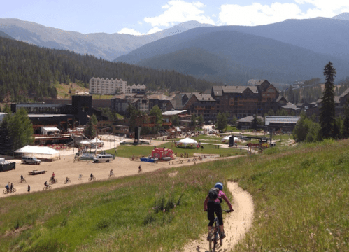 A cyclist rides on a dirt path with a mountain resort and festival area in the background, surrounded by lush greenery.