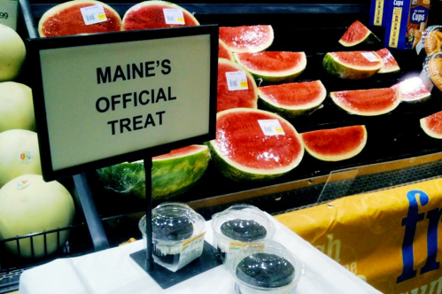 Sign reads "Maine's Official Treat" next to watermelons displayed in a grocery store.
