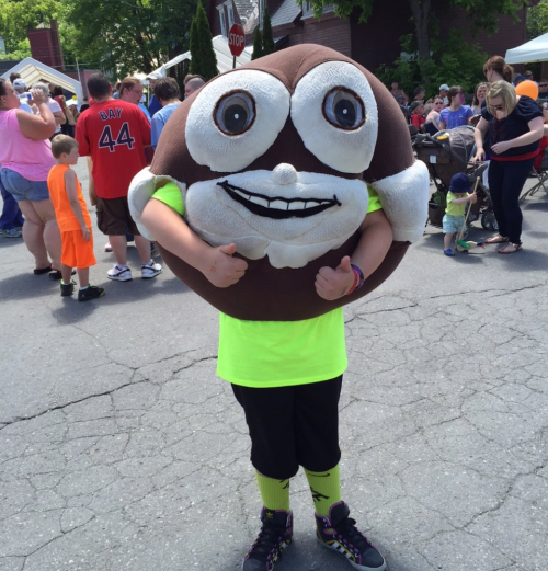 A person in a large chocolate cookie costume gives a thumbs up at a lively outdoor event with a crowd in the background.