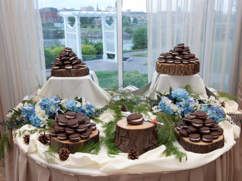 A dessert display featuring chocolate cookies on wooden stands, surrounded by blue flowers and pinecones, with a scenic view outside.