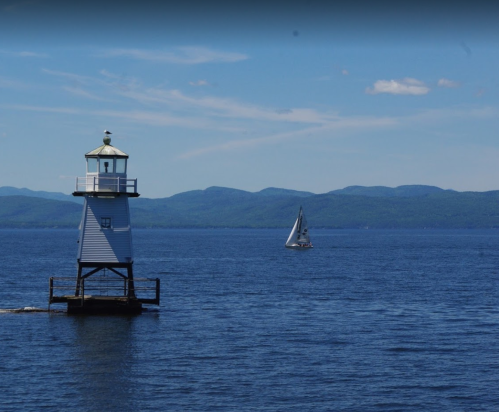 A lighthouse stands on a pier by the water, with a sailboat in the distance and mountains under a clear blue sky.