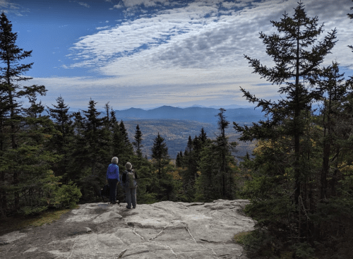 Two people stand on a rocky outcrop, overlooking a scenic mountain view surrounded by trees under a cloudy sky.