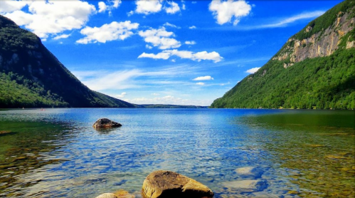 A serene lake surrounded by green mountains under a blue sky with fluffy clouds, featuring clear water and visible rocks.