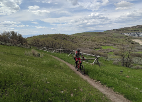A cyclist rides along a dirt path through green hills, with wooden fencing and a cloudy sky in the background.