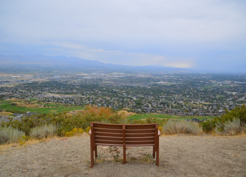 A wooden bench overlooks a vast valley with hills and a cloudy sky in the background.