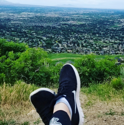 A person's feet in black sneakers resting on a grassy hill, overlooking a sprawling valley and distant mountains.