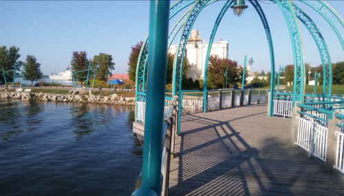A scenic view of a waterfront walkway with a teal arch, surrounded by trees and a calm water surface.