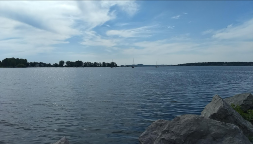 A serene view of a calm lake with distant sailboats and a clear blue sky, framed by rocky shoreline.