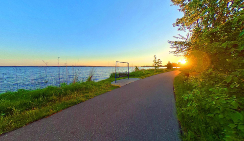 A serene lakeside path at sunset, with lush greenery and a swing set in the foreground.