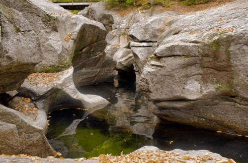 A serene rocky landscape with a calm pool of water surrounded by large boulders and autumn leaves.