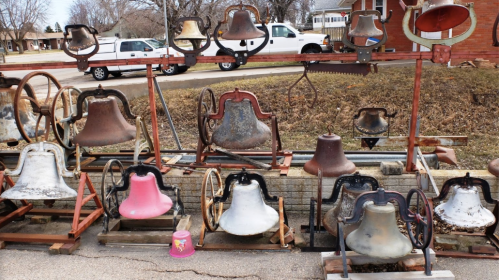 A collection of various old bells displayed on stands, with a mix of colors and sizes, set against a suburban backdrop.