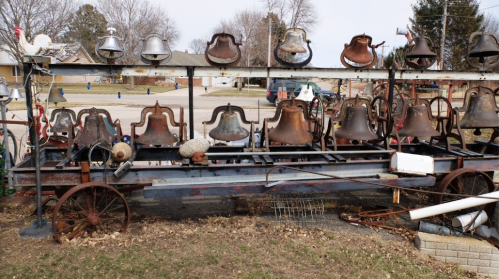 A collection of old, rusted bells displayed on a metal rack in a grassy area, with a few decorative items nearby.